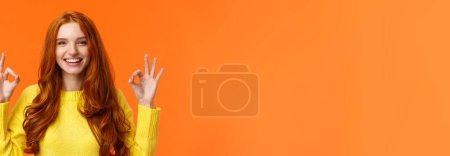 Mode, Shopping, Winterurlaubskonzept. attraktive fröhliche rothaarige Frau zeigt okay Gesten und Lächeln, alles in Ordnung, genehmigen oder wie Produkt, empfehlen Online-Shop, orangefarbener Hintergrund.
