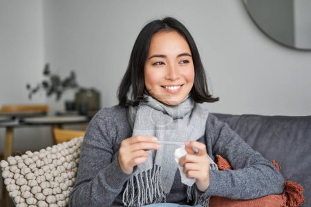 Foto de Mujer asiática sonriente sosteniendo el termómetro y mirando complacido, sintiéndose mejor después del frío, se deshizo de la fiebre, tiene temperatura normal. - Imagen libre de derechos