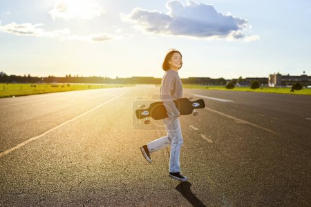 Téléchargez les photos : Asiatique fille avec skateboard debout sur la route pendant le coucher du soleil. Skater posant avec sa longue planche, pont de croiseur pendant l'entraînement. - en image libre de droit