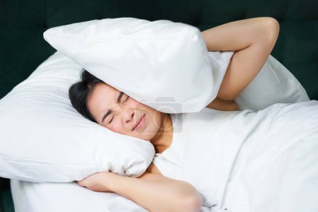 Téléchargez les photos : Triste fille avec insomnie. Jeune femme asiatique couchée au lit, couvrir ses oreilles avec oreiller, sommeil cant, partenaire snors, bruit fort la nuit la dérangeant. - en image libre de droit