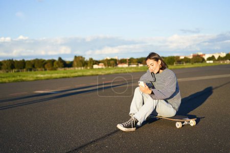 Téléchargez les photos : Portrait d'une jeune coréenne assise sur son skateboard sur la route, regardant son smartphone, bavardant sur une application mobile. - en image libre de droit