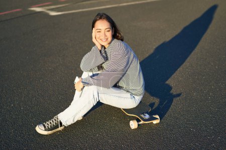 Téléchargez les photos : Portrait de femme asiatique assise sur skateboard, patinant sur son cruiser longboard, en utilisant l'application smartphone. - en image libre de droit