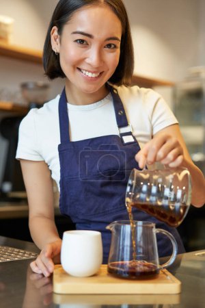 Téléchargez les photos : Plan vertical de sourire fille barista mignon, verser du café filtre, rendre l'ordonnance pour le client du café, portant tablier bleu. - en image libre de droit
