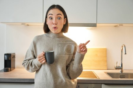 Téléchargez les photos : Portrait de sourire asiatique fille debout dans la cuisine, boire du café de tasse et pointant vers la bannière, montrant la publicité des agences de location. - en image libre de droit