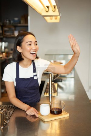 Téléchargez les photos : Souriant asiatique fille barista appelant le client, donner l'ordre dans le café, préparé mélange de café filtre, portant tablier derrière le comptoir. - en image libre de droit