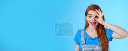 Photo for Dreamy curious attractive cheerful redhead woman surprised, look fascinated camera look through fingers okay sign, stand amused, gaze admiration blue background, check out promo. Copy space - Royalty Free Image