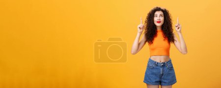 Photo for Woman wishing on shooting start believing in miracle looking and pointing hopefully up dreaming and praying in plan fullfillment standing in stylish urban cropped top over orange background. Copy - Royalty Free Image