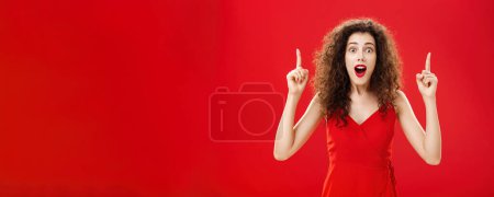 Photo for Woman stunned and impressed of huge sales. on clothes gasping and dropping jaw from amazement standing over red background in evening dress with make-up and curly hairstyle pointing up with raised - Royalty Free Image
