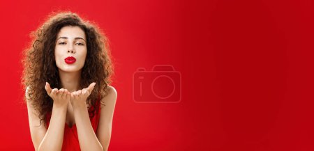 Téléchargez les photos : Femme récompense petit ami avec baiser vent passionné après rendez-vous romantique. Portrait de femme adulte chaude sensuelle en robe élégante rouge avec coiffure bouclée se penchant vers la caméra envoyant mwah flirt - en image libre de droit