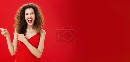 Photo for Woman feeling awesome on great party of friend pointing at entrance. Stylish carefree curly-haired female in red evening dress having fun near pool winking smiling joyfully and pointing left. - Royalty Free Image