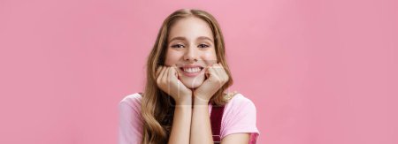 Lifestyle. Cute feminine and tender nice girl withtattoo and wavy hair leaning head on hands as if listening amusing story gazing with broad smile and admiration gaze being dreamy over pink background
