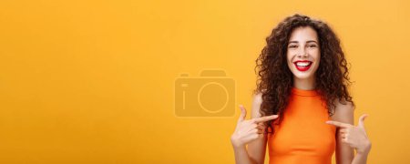 Foto de Foto en la cintura de la orgullosa y feliz mujer encantadora saliente con el pelo rizado y lápiz labial rojo apuntando a sí misma encantada y confiada hablando de su propio logro sobre fondo naranja. Copiar espacio - Imagen libre de derechos