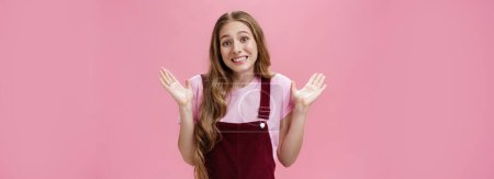 Sorry I have no idea. Silly unaware cute friendly-looking young female student in dungarees over t-shirt shrugging with palms raised in surrender smiling awkward as if being clueless over pink wall