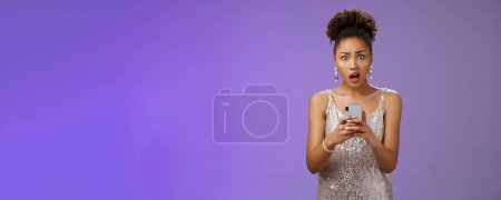 Photo for Shocked upset african-american woman in glittering stylish expensive dress drop jaw cringing disappointed receive terrible upsetting message holding smartphone look concerned blue background. - Royalty Free Image
