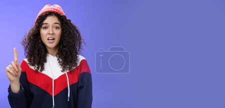 Photo for Rule number one do not mess with me babe. Portrait of swag and cool stylish young curly-haired woman in winter beanie showing index finger in prohibition or warning gesture over blue background. - Royalty Free Image