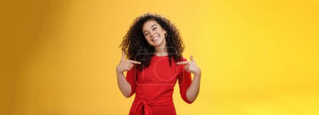 Lebensstil. Stolze und zufriedene ehrgeizige erfolgreiche Studentin im roten Kleid steht zufrieden lächelnd da und zeigt auf sich selbst, als prahle sie glücklich und froh über die gelbe Wand mit ihren eigenen Leistungen.