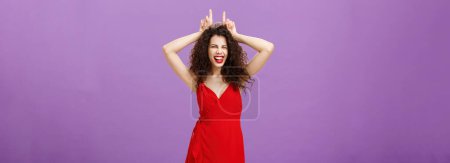 Photo for Devil lives inside lady. Daring stylish adult woman with curly hairstyle in red evening dress winking making confident and amused expression showing horns with index fingers on head, being stubborn. - Royalty Free Image