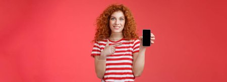 Photo for How about this app. Redhead curly friendly cute girl help friend find oufit online store hold smartphone pointing phone screen smiling broadly recommend social media photo filter, red background. - Royalty Free Image