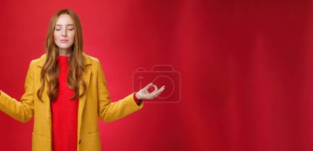 Foto de Chica mantener la calma liberando estrés con la meditación, posando en abrigo amarillo, cerrar los ojos y mirando aliviado como extender las manos de lado con gesto mudra, haciendo yoga sobre fondo rojo en pose de loto. - Imagen libre de derechos
