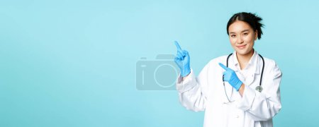 Photo for Smiling female doctor, physican in medical uniform and sterile gloves, pointing fingers left at promo, clinic logo, standing over blue background. - Royalty Free Image