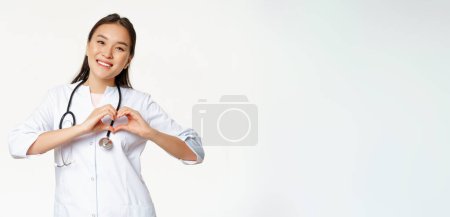 Photo for Smiling asian female doctor care for her patients, shows heart gesture and looking happy, standing in medical clinic uniform, white background. - Royalty Free Image
