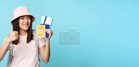 Téléchargez les photos : Fille voyageuse enthousiaste, touriste asiatique montre pouces levés et certificat de vaccin international, passeport de santé et billets d'avion, debout sur fond bleu. - en image libre de droit