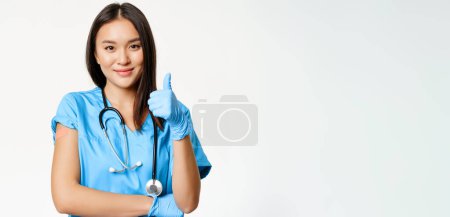 Lächelnde Krankenschwester, asiatische Ärztin im Peeling, Daumen hoch und geimpfter Arm mit medizinischem Pflaster, empfiehlt Impfung ab Covid-19, weißer Hintergrund.