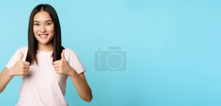 Téléchargez les photos : Fille asiatique enthousiaste montrant pouces vers le haut, comme et approuver, souriant satisfait, debout sur fond bleu. Espace de copie - en image libre de droit
