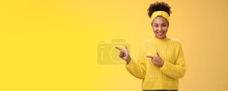 Seguro de buena apariencia milenaria femenina afroamericana mujer diadema suéter mirada asertiva apuntando dedos índice izquierdo que te va a gustar sonrisa, de pie fondo amarillo.
