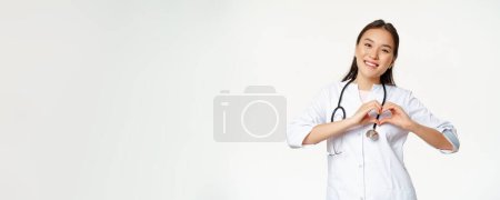 Photo for Smiling asian female doctor care for her patients, shows heart gesture and looking happy, standing in medical clinic uniform, white background. - Royalty Free Image