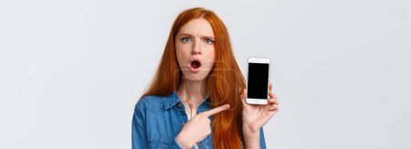 Téléchargez les photos : Close-up portrait angry and pissed-off redhead girl showing photos from social media ex-boyfriend complaining to friend, pointing finger smartphone display and frowning upset, white background. - en image libre de droit