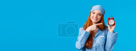 Dont forget wake up on time. Cheerful and cute relaxed feminine redhead woman in pyjama and sleep mask, holding red clock, pointing at alarm as set up before sleep, standing blue background.