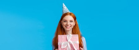 Photo for Waist-up portrait cheerful happy, pretty redhead, ginger girl celebrating birthday, receive cute pink wrapped present, wearing b-day cap and smiling, having fun at party, blue background. - Royalty Free Image