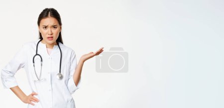 Téléchargez les photos : Confondu asiatique femme médecin cant comprendre patient, regardant perplexe et ennuyé, levant la main vers le haut questionné pose, debout en uniforme médical sur fond blanc. - en image libre de droit