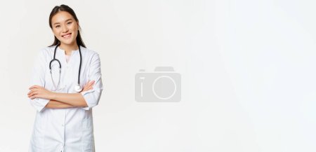 Photo for Asian female doctor, physician in medical uniform with stethoscope, cross arms on chest, smiling and looking like professional, white background. - Royalty Free Image