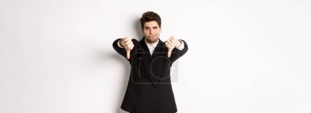 Photo for Portrait of skeptical and disappointed man in black suit, frowning upset, showing thumbs-down, dislike something bad, standing over white background. - Royalty Free Image