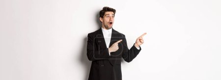 Photo for Impressed handsome man in party suit, looking at new year promo offer and pointing fingers left at banner, standing over white background. - Royalty Free Image
