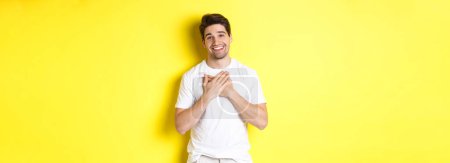 Téléchargez les photos : Image d'un beau garçon reconnaissant en t-shirt blanc, tenant la main sur le cœur et souriant heureux, exprimer sa gratitude, remercier pour quelque chose, debout sur fond jaune. - en image libre de droit