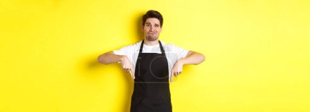 Photo for Skeptical barista in black apron pointing fingers down at bad product, looking displeased and unamused, standing over yellow background. - Royalty Free Image