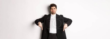 Photo for Portrait of skeptical and unamused bearded man, pointing fingers down with disappointed face, standing in suit and complaining. - Royalty Free Image