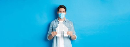 Photo for Concept of coronavirus, quarantine and social distancing. Young man searching apartment, showing house paper model, wearing medical mask, renting or buying propery, blue background. - Royalty Free Image