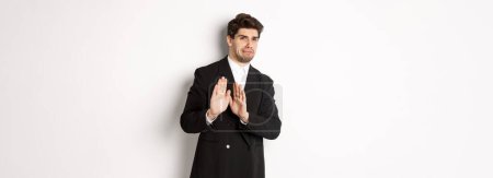 Photo for Image of stylish handsome man in black suit, cringe from something embarrassing, declining offer and showing stop gesture, standing against white background. - Royalty Free Image
