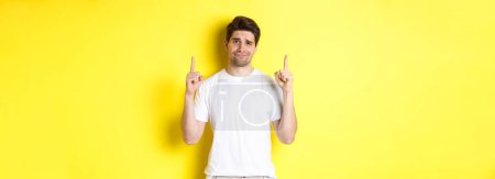 Photo for Unamused handsome guy frowning, pointing fingers up at something bad, standing skeptical against yellow background. - Royalty Free Image