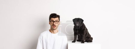 Bild einer Hipster-Hundebesitzerin, die neben einem niedlichen schwarzen Mops sitzt und verwirrt in die Kamera starrt, weißer Hintergrund.