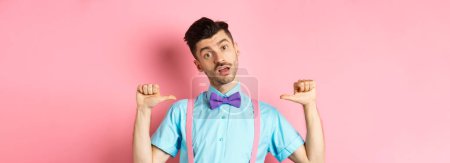 Photo for Sassy and confident young man in suspenders and bow-tie, pointing at himself and looking like professional, standing over pink background. - Royalty Free Image