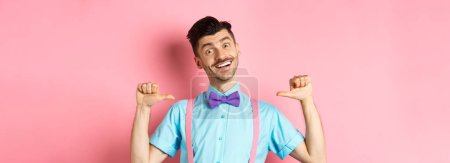 Photo for Cheerful smiling man with moustache and bow-tie, pointing at himself and self-promoting, standing happy over pink background in suspenders. - Royalty Free Image