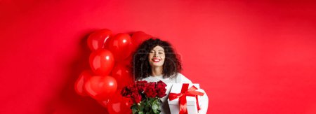 Photo for Smiling happy woman holding box with gift and red roses from boyfriend, celebrating Valentines day, standing near romantic hearts balloons, standing over studio background. - Royalty Free Image