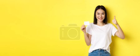 Joven mujer asiática en casual camiseta blanca mostrando tarjeta de crédito de plástico y el pulgar hacia arriba gesto, aprobar y recomendar, sonriendo a la cámara, fondo amarillo.
