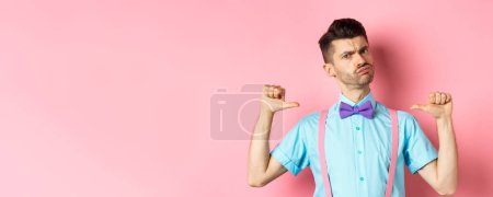 Photo for Serious and funny young man trying look cool, pointing at himself to self-promote, being a professional, standing over pink background. - Royalty Free Image