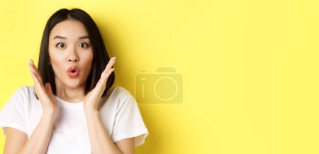 Photo for Close up of surprised asian girl say wow, stare at camera amzed with hands near face, standing over yellow background. - Royalty Free Image